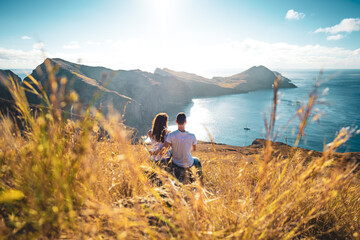 Fototapeta na wymiar Back view of a tourist couple sitting on a field with coastal landscape of Madeira Island in the Atlantic Ocean in the morning. São Lourenço, Madeira Island, Portugal, Europe.