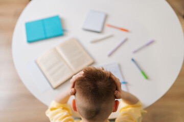 High angle back view of exhausted pupil boy tired from studying holding head head with hands sitting at desk with paper copybook, looking down. Frustrated child schoolboy doing homework at home.