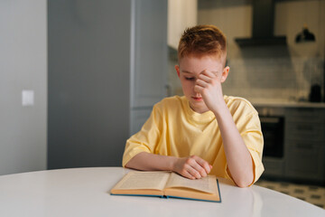 Portrait of focused cute little student boy reading paper book following lines with finger along...
