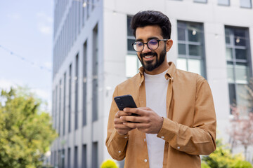Successful Indian young businessman outside office building walking in daytime, man holding phone...