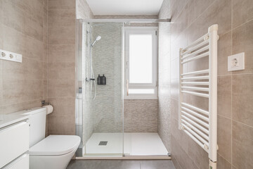 Naklejka na ściany i meble Comfortable bathroom with a toilet bowl and a shower cabin with tiles in beige tones and a window for natural light. The concept of a bathroom in a hotel or apartment after renovation