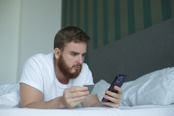 Young happy man doing online shopping with smartphone, lay in bed in bedroom at home, hold in hand credit bank card and cell mobile phone and smile