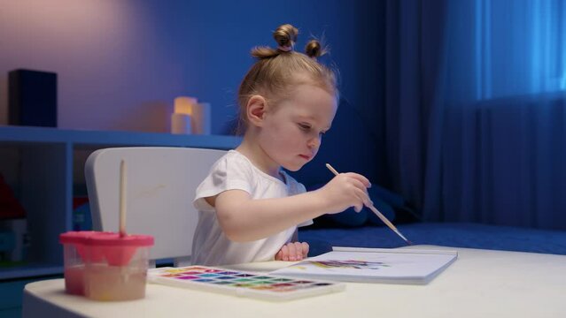 Child paints with watercolor paints sitting at table in evening. Cute two-year-old blonde girl paints multicolored paints on white paper sitting at home at table in evening.