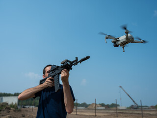 A man aims to shoot a rifle at a flying drone against a blue sky. 