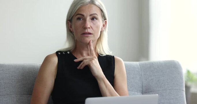 Thoughtful busy mature freelance woman working with laptop on home couch, thinking over project ideas, looking away, touching chin, making decision, typing, turning to screen