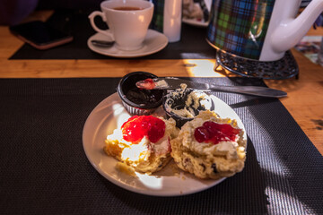 Fine Scottish food: fruit scone warm with whipped butter and strawberry jam plated in bars of sunlight on a cafe table tea cup and pot in background. Shot in Comrie strathearn perthshire scotland