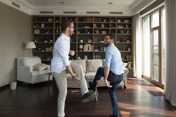 Two happy cheerful young adult twin brothers having fun at home, dancing together in living room,...