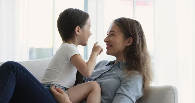 Cheerful adorable sweet kid sitting on mothers lap on home couch, chatting to mom, playing game, giving high five, kissing mommy. Young mum having fun with child, speaking, laughing