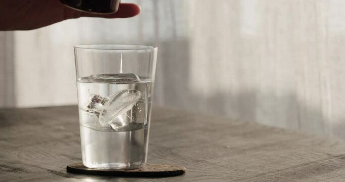 Slow motion man pour espresso into tonic with clear ice cube in tumbler glass on black oak table