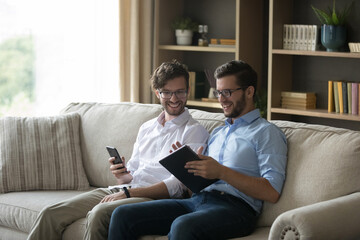 Obraz premium Happy twin brother men discussing successful business project at home, using watching online startup on tablet, using digital devices on conversation, sitting on home couch, talking, laughing