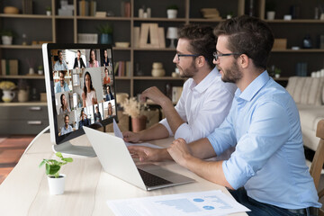 Focused twin family business partners speaking on online conference video call, using large desktop monitor with head shots, talking to diverse freelance team of colleagues, discussing teamwork - obrazy, fototapety, plakaty