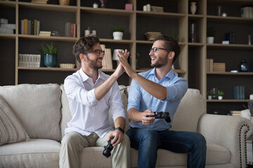 Happy young adult twin brothers playing video game at home, enjoying virtual cyber competition,...