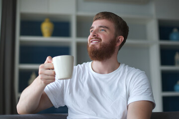 Positive young european beard looking man having a hot beverage and drink from mug in living room, at home