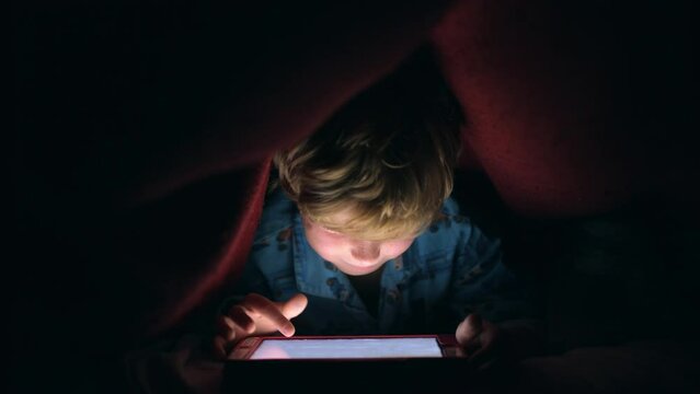 Night, light and tablet with child with blanket after bedtime for internet, movie and technology for streaming service. Relax, learning and digital with boy in bedroom hiding and playing online games - Powered by Adobe