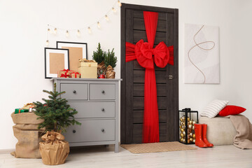 Beautiful fir tree and sack with Christmas gifts near chest of drawers and wooden door decorated with red bow in room
