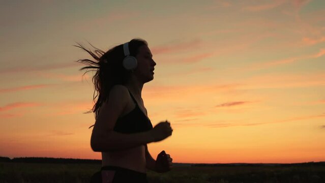 Active young woman runs in summer in park at sunset, listens to music on headphones. Silhouette, beautiful girl in musical headphones, is engaged in fitness, runs in evening. Training run. Teen runner