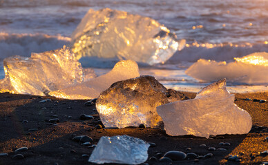 Pure glacier ice shining at sunset. Black volcanic sand at ocean coast. Pure ocean waves crash golden icebergs in Iceland. Diamond beach - famous tourist location in north Europe country.