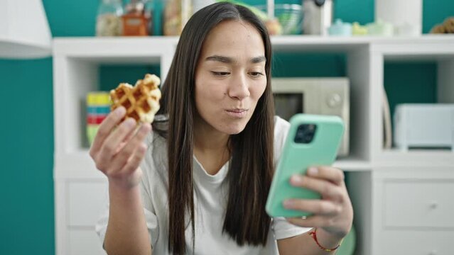 Young beautiful hispanic woman eating waffle using smartphone at dinning room