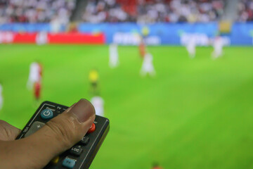 A man is pointing a black remote control at an unfocused tv while watching football