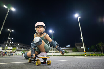 asian child exercise or kid girl skater playing skateboard or surf skate and squatting down in skatepark or extreme sports park at night to sitting on board and wears helmet knee guard for body safety