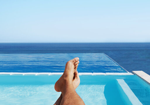 POV, feet and man relax at a pool for travel and summer vacation at the sea by hotel and water. Barefoot, calm and traveling person outdoor at poolside holiday or rest at luxury resort in Maldives