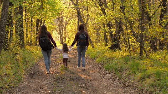 Happy family life concept. young women walk through forest with small child with backpacks their backs. walk woods summer weekend sunset. little girl holding hands parents walking along path summer