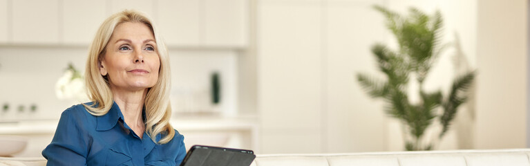 Relaxed mature lady in casual wear looking away, using tablet pc while sitting on a couch in modern...