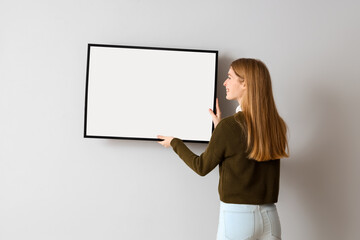 Young woman hanging blank frame on light wall