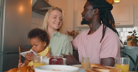 Lovely couple flirting at home while having breakfast in slow motion. Handsome husband kissing wife, adorable little daughter sits on mom's knees, using gadget, playing games, watching cartoon - Powered by Adobe
