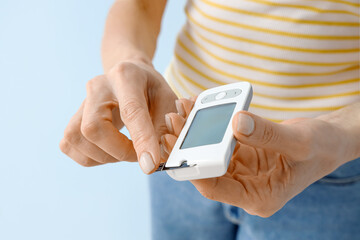 Mature diabetic woman measuring blood sugar level with glucometer on blue background, closeup