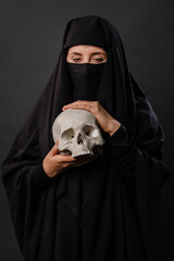 Portrait of a young, adult woman in a black burqa with a hidden face, with a skull in hands...