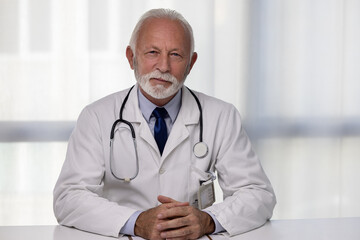 Portrait of senior male doctor, sitting by the table and looking at a camera. Medical director professional of hospital wearing uniform and showing confidence - obrazy, fototapety, plakaty