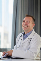 Portrait of friendly doctor man sitting by the desk, looking away and laughing, being relaxed, cheerful, feels confident and successful