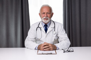 Portrait of senior male doctor, sitting by the table and looking at a camera. Medical director professional of hospital wearing uniform and showing confidence
