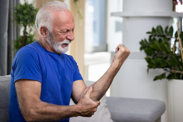 Elderly man holding his elbow, suffering from arthritis pain in hand, sitting on sofa at home - 602222402