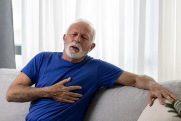 Senior man holding his chest and feeling pain suffering from heart attack sitting on sofa at home
