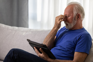 Senior man using digital tablet computer at home and having eyestrain or suffering from astigmatism...
