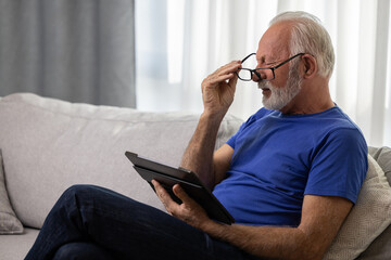 Senior man with glasses using digital tablet computer at home and having eyestrain or suffering from astigmatism and bad vision. Eyesight problems concept.