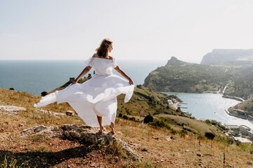 Happy woman in a white dress and hat stands on a rocky cliff above the sea, with the beautiful silhouette of hills in thick fog in the background.