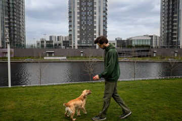 A young man walks his dog in the park on a cloudy morning. A bearded man gives his dog a small red ball during a morning walk.