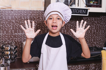 Happy excited cute Asian kid in chef uniform showing flour hands at camera, smiling, at the...