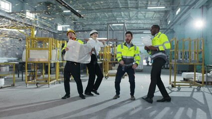 Multicultural engineers dancing at modern manufacturing factory and clapping hands. Employees in safety hard hats celebrating succsess at background of welding sparks flying.