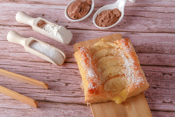 top view apple pie with grated coconut on a wooden board spoons with chocolate, cocoa flour and sugar and wooden tongs