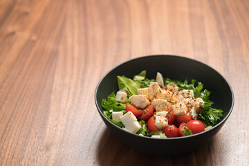 Healthy light salad with cherry tomatoes, mozzarella and frisee in black bowl on walnut surface