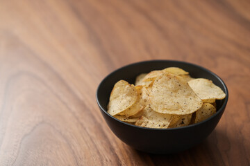 Organic potato chips with black pepper in black ceramic bowl on walnut wood background with copy space