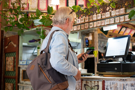 Senior white-haired man pays at the coffee shop checkout using the application on his mobile phone. Concept of modernity and online electronic payments - Powered by Adobe
