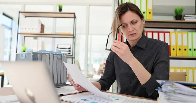 Tired woman manager takes off glasses looking at papers sitting at table with laptop. Female office employee checks up commercial data at workplace slow motion