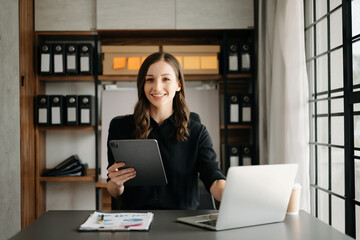 Confident business expert attractive smiling young woman typing laptop ang holding digital tablet on desk in office.