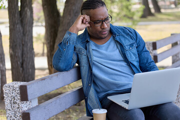 Fototapeta premium Modern businessman carefully checks reports of employees sitting on bench on summer day. Stylish man in denim shirt performs work in comfortable place outside the office thanks to modern technology