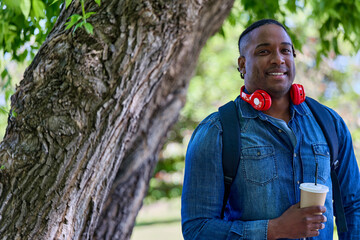 Cute African student relaxes in park with glass of coffee in between classes. Walking through park, a dark-skinned student listens to an audio book on a given topic with the help of modern headphones